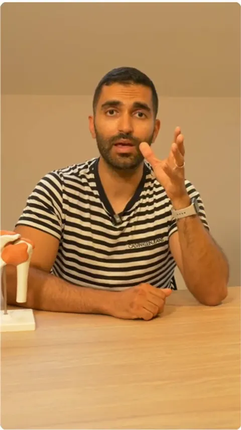 Aziz Youssef explique la capsulite face caméra, un modèle d'épaule posé sur son bureau, dans la vidéo pédagogique qu'il joint à sa prescription